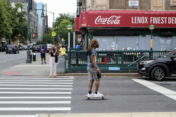 a guy on a skateboard rides past a subway stop in Harlem